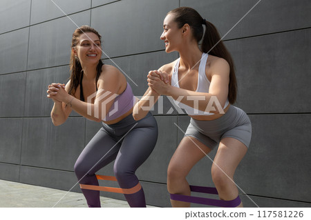 Two smiling young women doing squats with fitness band Two smiling young women doing squats with fitness band 117581226