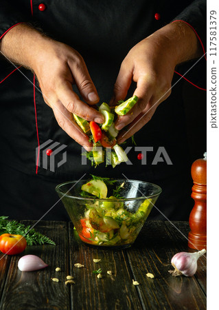 The cook prepares a vegetable mix for lunch. We prepare a salad with fresh vegetables and spices on the kitchen table. The chef hands mix the chopped vegetables in a bowl 117581379