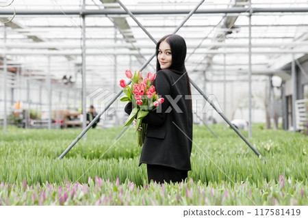 Young woman standing in large greenhouse and holding a blooming tulips in her hands Young woman standing in large greenhouse and holding a blooming tulips in her hands 117581419