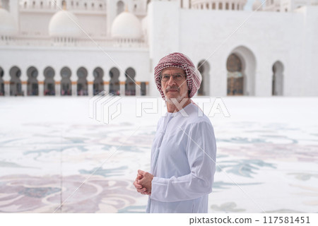 Man in traditional attire poses outside grand mosque with domes and arches. 117581451
