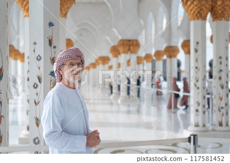 Man in traditional attire stands inside ornate mosque with columns behind. 117581452