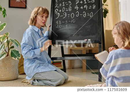 Young woman sitting on floor and holding white chalk while explaining numbers to her daughter 117581609