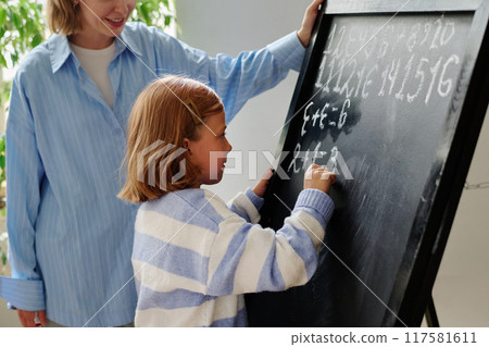 Little girl with bob haircut standing next to black board and doing sums while he mom praising her Little girl with bob haircut standing next to black board and doing sums while he mom praising her 117581611