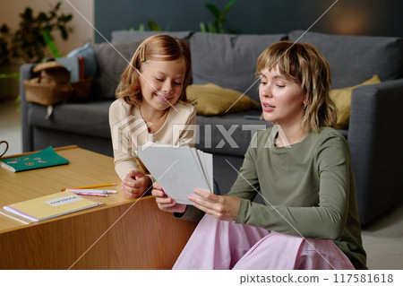 Young mom sitting on floor and holding sketches while discussing them with her lovely girl Young mom sitting on floor and holding sketches while discussing them with her lovely girl 117581618