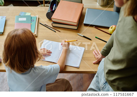 High angle shot of daughter sitting at wooden table and doing sums while her mother watching her 117581667