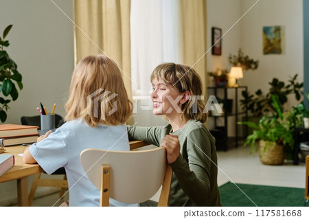 Back view of little girl sitting at wooden table while her mom sitting on floor next to her 117581668