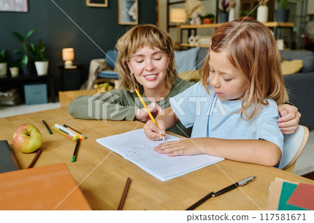 Young mom hugging her daughter while she sitting at wooden table and doing math 117581671