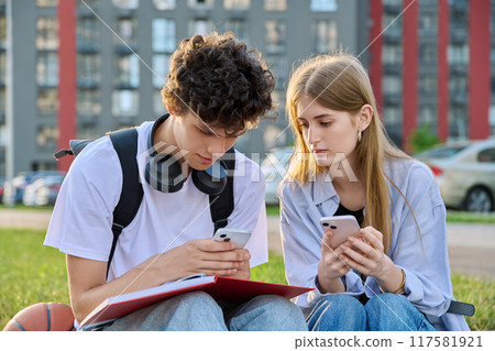 Teenage college students guy and girl talking, using smartphone, sitting outdoor Teenage college students guy and girl talking, using smartphone, sitting outdoor 117581921