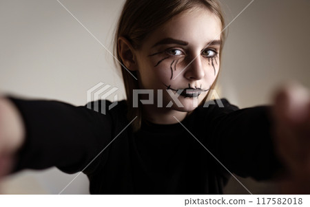 Little girl with spooky Halloween makeup looking at camera and taking selfie. Creepy kid's portrait in black and white. 117582018