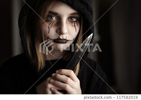 Spooky little girl holding knife and looking at blade. Scary child kid in black and Halloween dark makeup Spooky little girl holding knife and looking at blade. Scary child kid in black and Halloween dark makeup 117582019