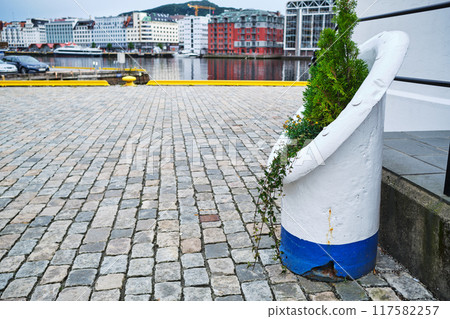 Waterfront scene in Bergen, Norway, featuring cobblestone paving, urban architecture, and a decorative planter made from a repurposed marine bollard 117582257