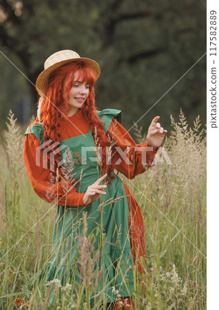A shepherdess woman with red hair and a green dress sits in a field of flowers. 117582889