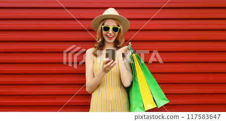 Beautiful happy cheerful woman looking at phone with shopping bag in summer hat on red background Beautiful happy cheerful woman looking at phone with shopping bag in summer hat on red background 117583647
