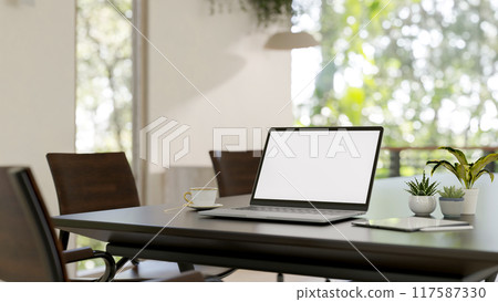 A laptop with a white-screen mockup on a hardwood meeting table in a bright modern meeting room. 117587330
