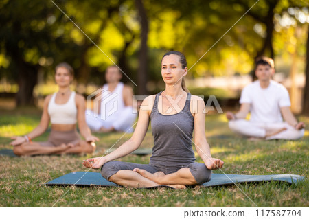 Woman meditating in Padmasana pose during group yoga in park 117587704