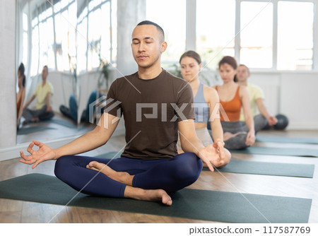 Positive man in sports clothes having yoga class in group sitting in lotus pose in yoga studio Positive man in sports clothes having yoga class in group sitting in lotus pose in yoga studio 117587769