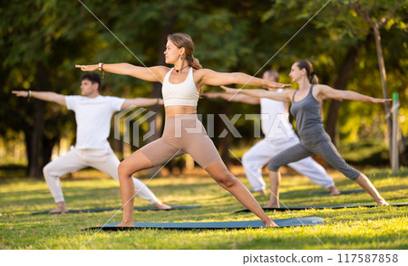 Girl performing Warrior pose during group yoga session in park Girl performing Warrior pose during group yoga session in park 117587858