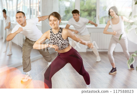 Happy young girl in sportswear with group active people practising twist or charleston dancing in dance hall 117588023