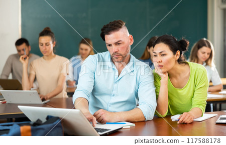 Two students studying is on a laptop, discussing something Two students studying is on a laptop, discussing something 117588178