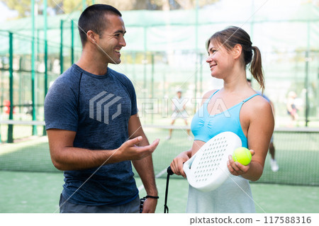 Portrait of two woman and man in sportswear on outdoor court with paddle racket and ball in hands after match, talking friendly Portrait of two woman and man in sportswear on outdoor court with paddle racket and ball in hands after match, talking friendly 117588316