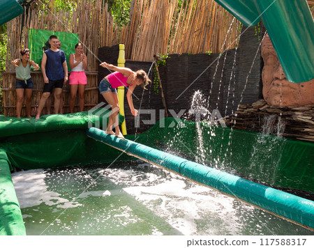 Cheerful woman trying to walk on inflatable log above pool of water Cheerful woman trying to walk on inflatable log above pool of water 117588317