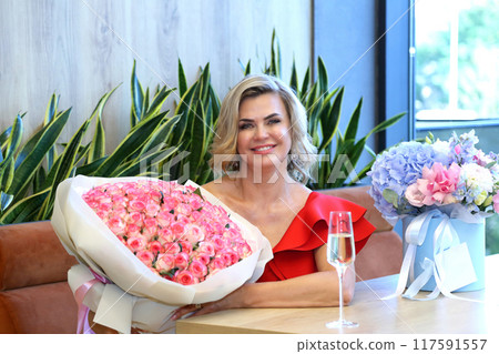 Beautiful woman in a red dress. Beautiful mature woman with champagne. A gorgeous adult beauty in a red dress with flowers and champagne. Gorgeous adult beauty. 117591557