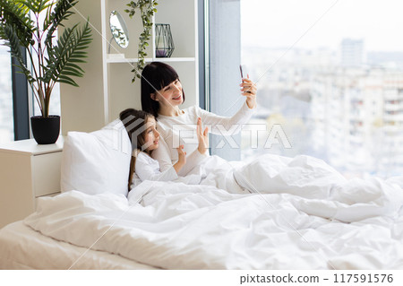 Mom and daughter lying in bed having video call on phone 117591576
