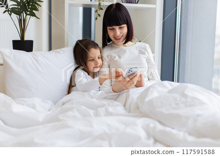 Mom and daughter lying in bed using smartphone together Mom and daughter lying in bed using smartphone together 117591588