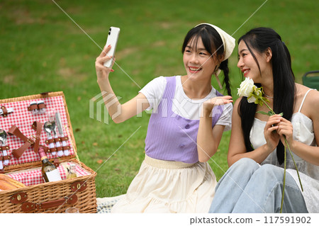 Two cheerful asian women sitting on a grassy field, taking a picture with smartphone during picnic Two cheerful asian women sitting on a grassy field, taking a picture with smartphone during picnic 117591902