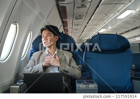 Young businessman looking out the window with hand resting on his chest, experiencing aerophobia or fear of flying 117591914