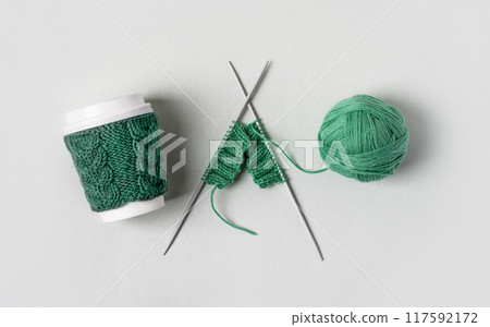 Cup for takeaway coffee with knitted green cover and knitted wool with knitting needles on gray background. Concept of takeaway coffee, walking, energetic life, hobbies needlework 117592172