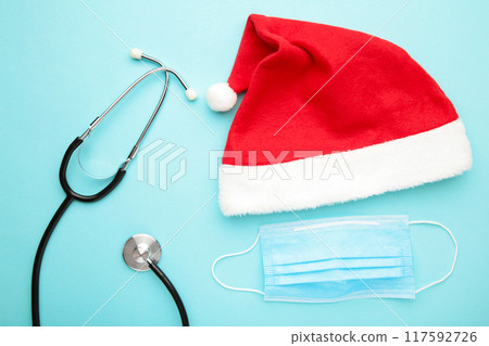 Medicine christmas new year concept during coronavirus covid time. Top view of medical stethoscope and protective mask with santa hat on blue background. Medicine christmas new year concept during coronavirus covid time. Top view of medical stethoscope and protective mask with santa hat on blue background. 117592726