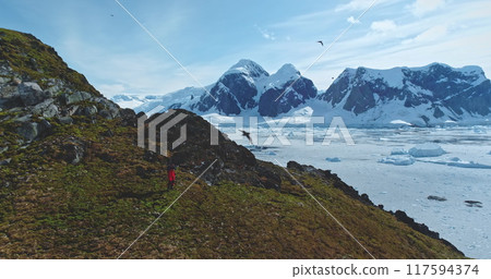 Tourist explore Antarctica wild nature landscape. Man trekking take photo of mountains green rock hill wildlife. Frozen ocean, icebergs snow covered island in background. Polar winter seagulls. Aerial 117594374