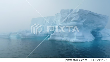 Large iceberg floating in polar ocean in foggy mist day in Antarctica. Tabular huge high ice glacier melting ice cave at wild nature environment. Arctic winter landscape at global warming problem. 117594423