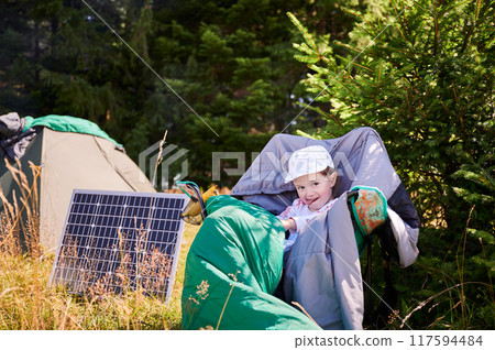 Young child playing near tourist tent with photovoltaic solar panel in summer. On background of dense forest. Integration of renewable energy in outdoor camping activities. Young child playing near tourist tent with photovoltaic solar panel in summer. On background of dense forest. Integration of renewable energy in outdoor camping activities. 117594484