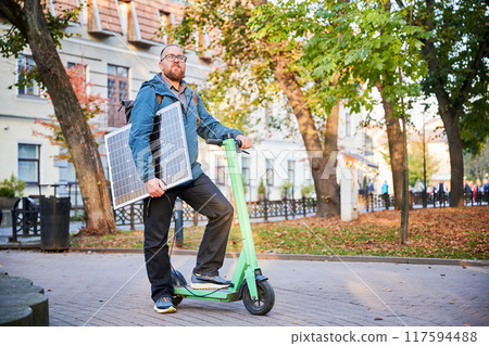 Man rides electric scooter while carrying solar panel. Integration of solar power as sustainable energy source for charging electric scooters, promoting eco-friendly urban transportation. Man rides electric scooter while carrying solar panel. Integration of solar power as sustainable energy source for charging electric scooters, promoting eco-friendly urban transportation. 117594488