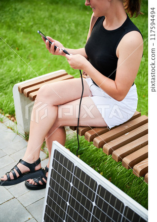 Woman connecting smartphone to photovoltaic solar panel. Cropped view. Integration of sustainable renewable energy into everyday life, demonstrating practical use of solar power for charging devices. 117594494