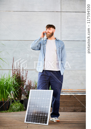 Bearded man talks on smartphone connected to photovoltaic solar panel. Integration of sustainable renewable energy into everyday life, demonstrating practical use of solar power for charging devices. Bearded man talks on smartphone connected to photovoltaic solar panel. Integration of sustainable renewable energy into everyday life, demonstrating practical use of solar power for charging devices. 117594500