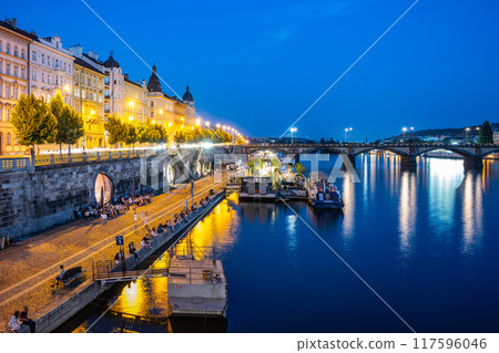 The Vltava River in Prague is bustling with activity at night. People are gathered on the embankment, enjoying the cool evening air. The city lights reflect off the water, creating a beautiful and The Vltava River in Prague is bustling with activity at night. People are gathered on the embankment, enjoying the cool evening air. The city lights reflect off the water, creating a beautiful and 117596046