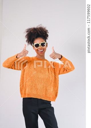 Smiling African American woman in orange sweater and heart-shaped sunglasses, giving a thumbs up on a light background. 117599602