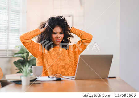 African American woman feeling stressed and holding her head in frustration while working on a laptop at home. 117599669