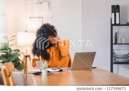 African American woman massaging her neck, indicating discomfort or strain while working on a laptop at home. 117599673