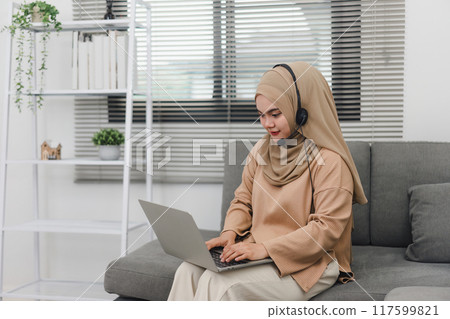 Muslim Business Woman wearing a hijab and headset works remotely on her laptop from a cozy home office. 117599821