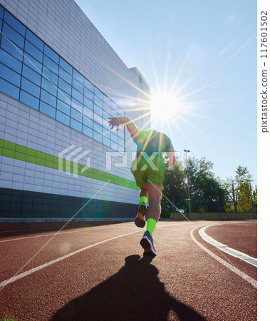 Runner charges ahead, silhouetted by bright sun, his green attire glowing as rays burst around him, symbolizing energy and determination. 117601502