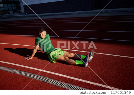 After tough run, athlete sits on the track, deep in thought, his green outfit contrasting sharply with vibrant red surface beneath him. 117601549