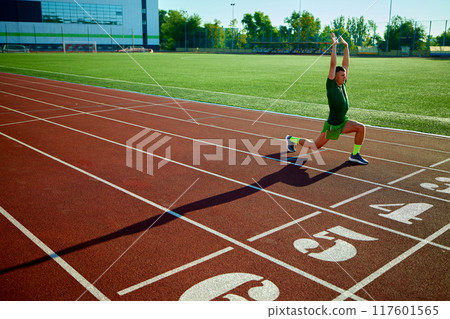 Young fit man, athlete stretches at starting line, preparing for race on sunlit track, with lush green field in background. 117601565