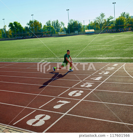 Sprinter, athletic man in green gear warms up with lunge at starting line, under bright morning sun on empty track. 117601566