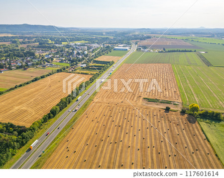 Aerial perspective showing a highway bordered by fields of hay bales, with rural landscape stretching into the distance. Aerial perspective showing a highway bordered by fields of hay bales, with rural landscape stretching into the distance. 117601614