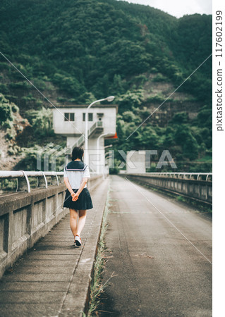 The back view of a girl in a sailor uniform walking along the road on a bridge 117602199