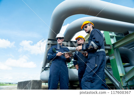 Side view of engineer or factory technician worker team stand under petrochemical gas pipeline with one also point to the pipeline in workplace. Side view of engineer or factory technician worker team stand under petrochemical gas pipeline with one also point to the pipeline in workplace. 117608367